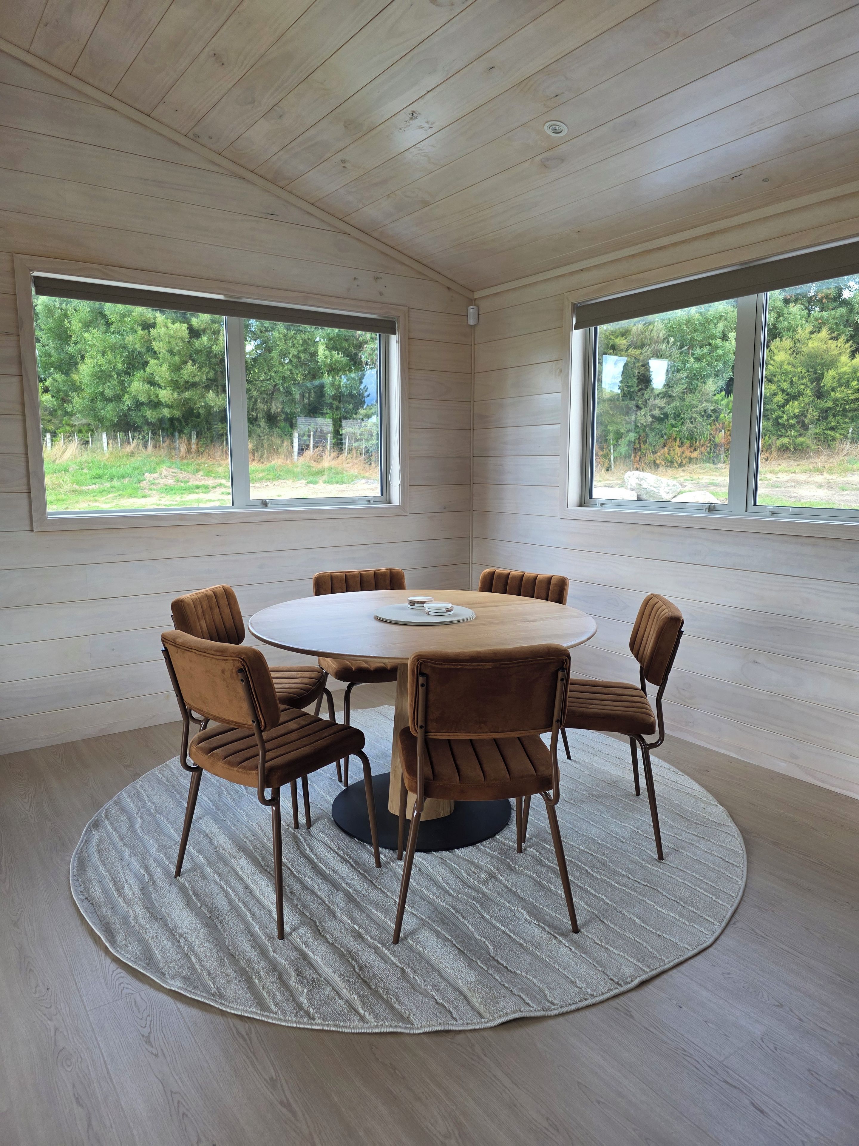 A room with a round wooden table surrounded by six brown chairs, large windows, and a view of greenery outside.