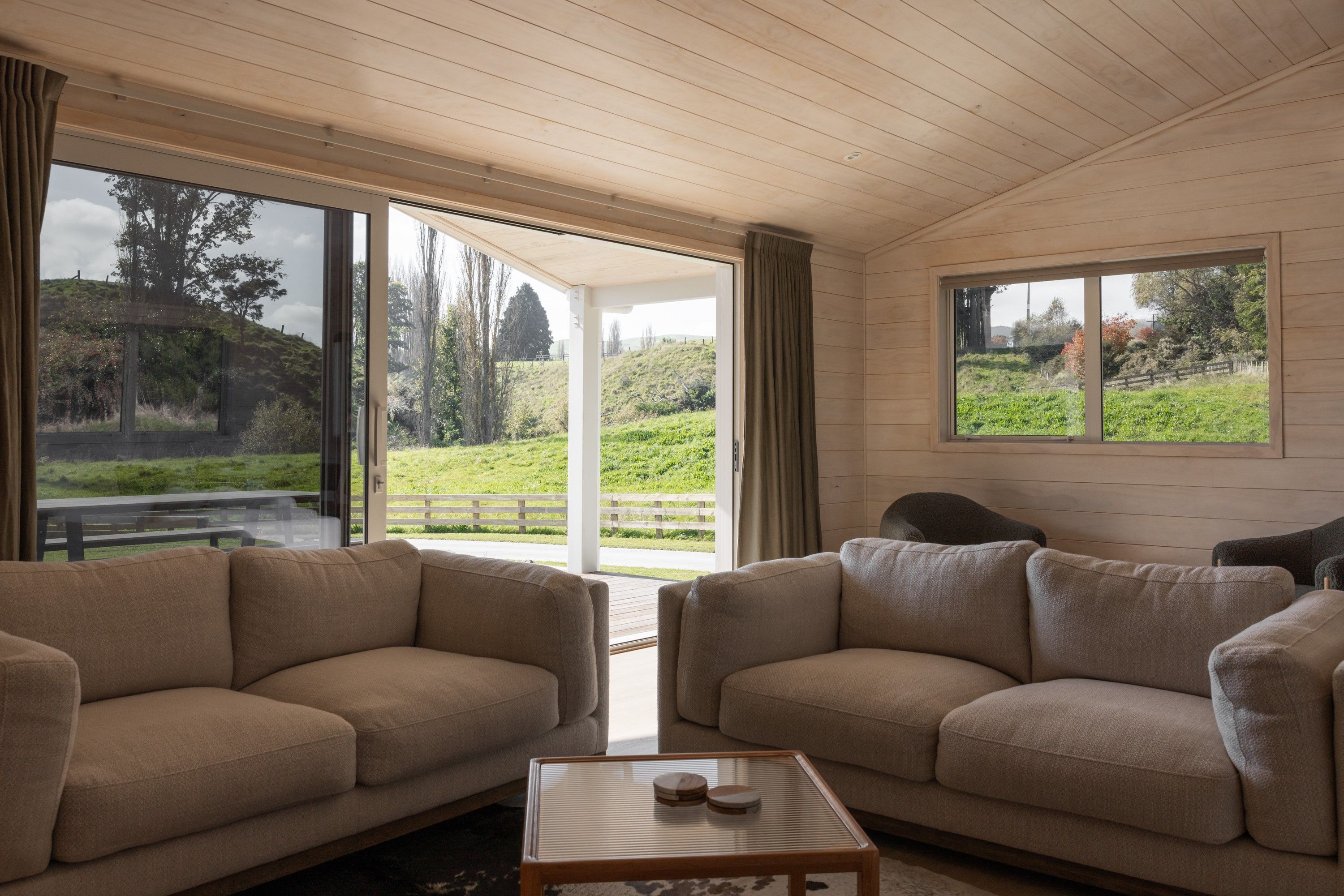 A room with two beige sofas, a glass coffee table, and a view of a green landscape through large windows.