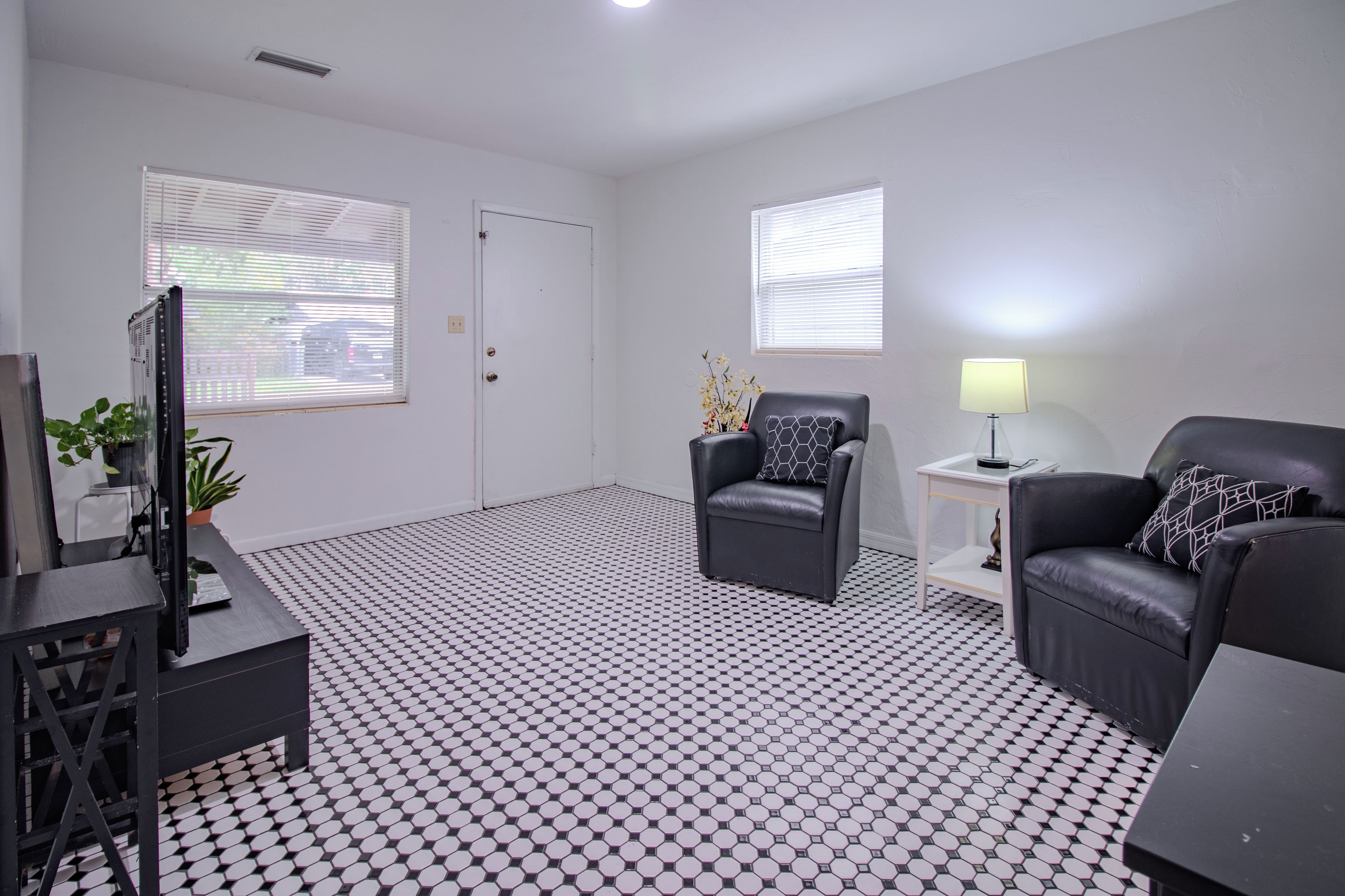 A modern living room with a black and white checkered floor, two black leather sofas with patterned cushions, a small side table with a lamp, and a television on a stand.
