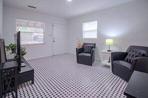 Ein modernes Wohnzimmer mit einem schwarz-weißen Schachbrettmuster-Boden, zwei schwarzen Ledersofas mit gemusterten Kissen, einem kleinen Beistelltisch mit Lampe sowie einem Fernseher auf einem Lowboard.