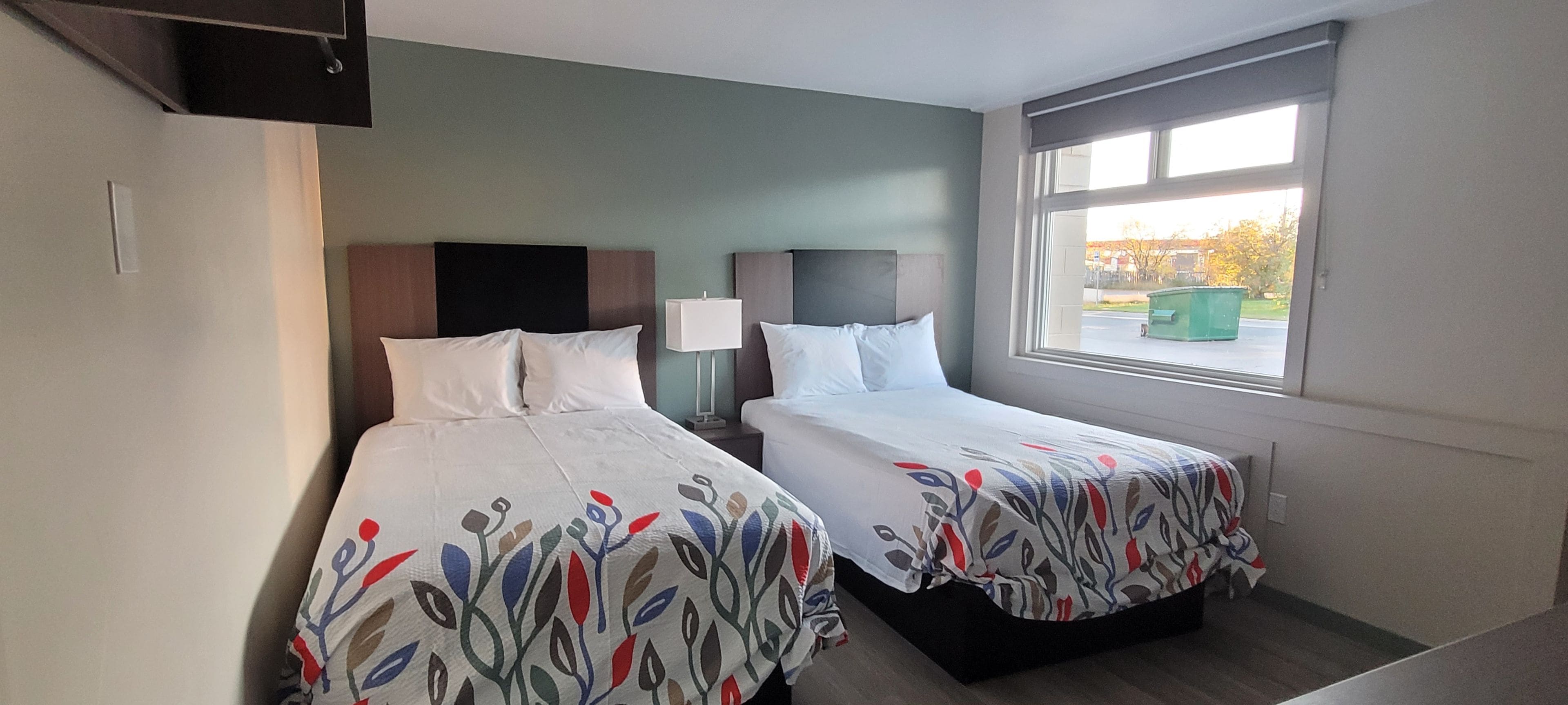 Ein Hotelzimmer mit zwei Betten, einem Fenster mit Blick nach draußen und einem an der Wand befestigten Flachbildfernseher.