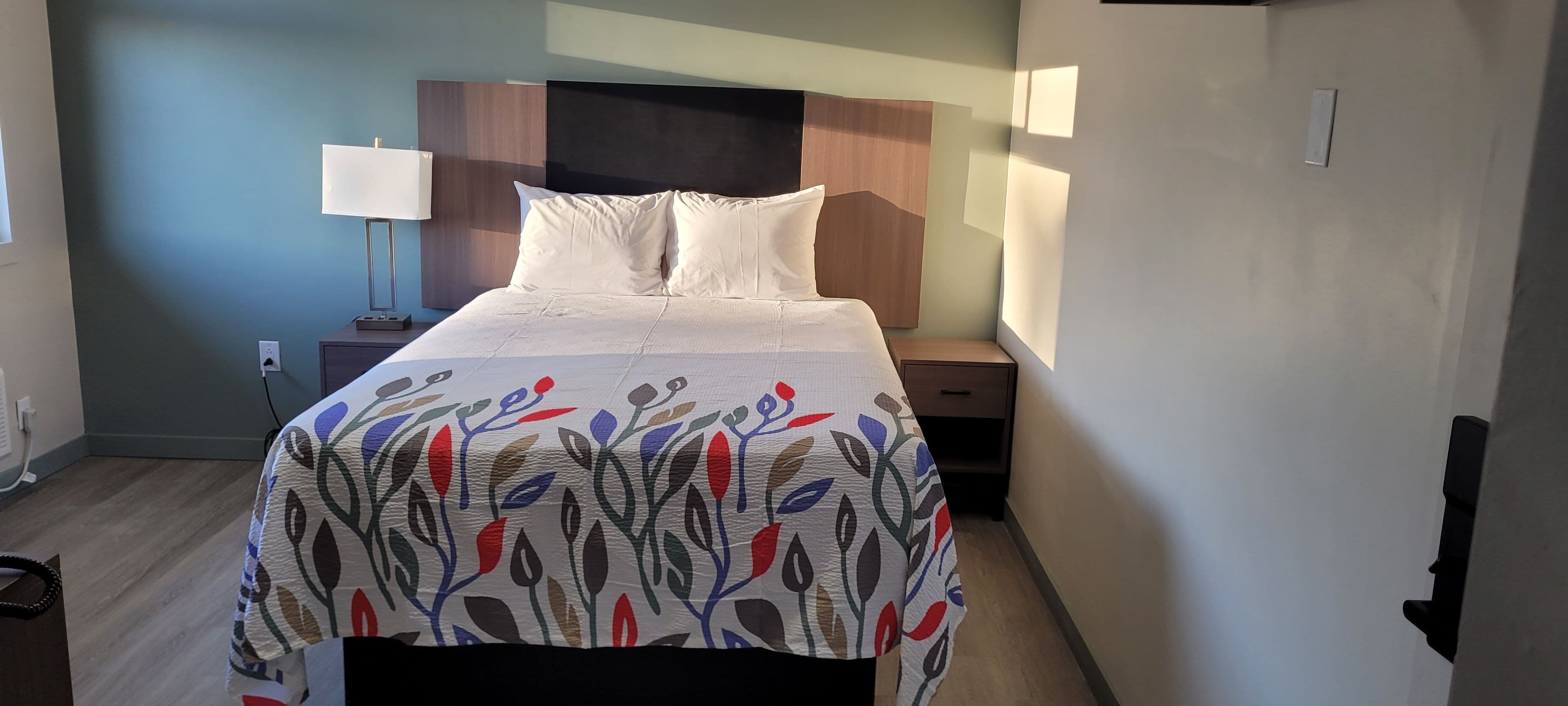Un lit bien fait, recouvert d’une couvre-lit à motifs floraux, avec une lampe de chevet et une table de nuit également dotée d’une lampe.