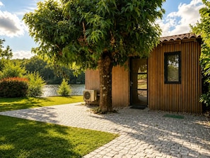 A wooden cabin with a door and window, surrounded by a paved walkway and lush greenery.