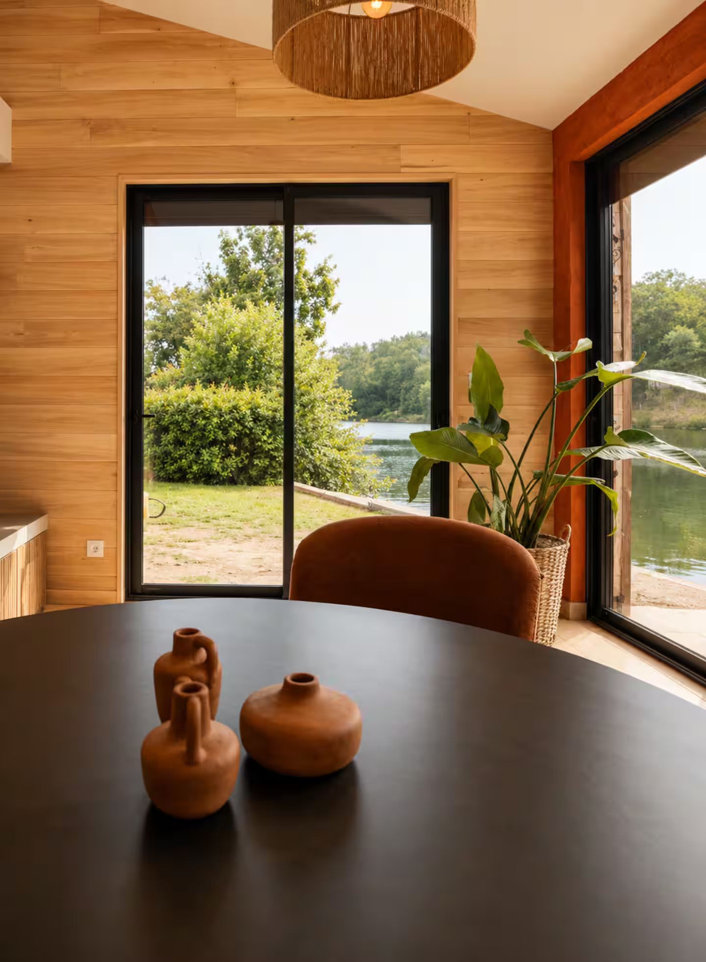 Un intérieur moderne avec un mur en bois, une grande fenêtre donnant sur un paysage verdoyant et une étendue d’eau, une table ronde agrémentée de deux objets en céramique et un pot de fleurs en fibres tressées.
