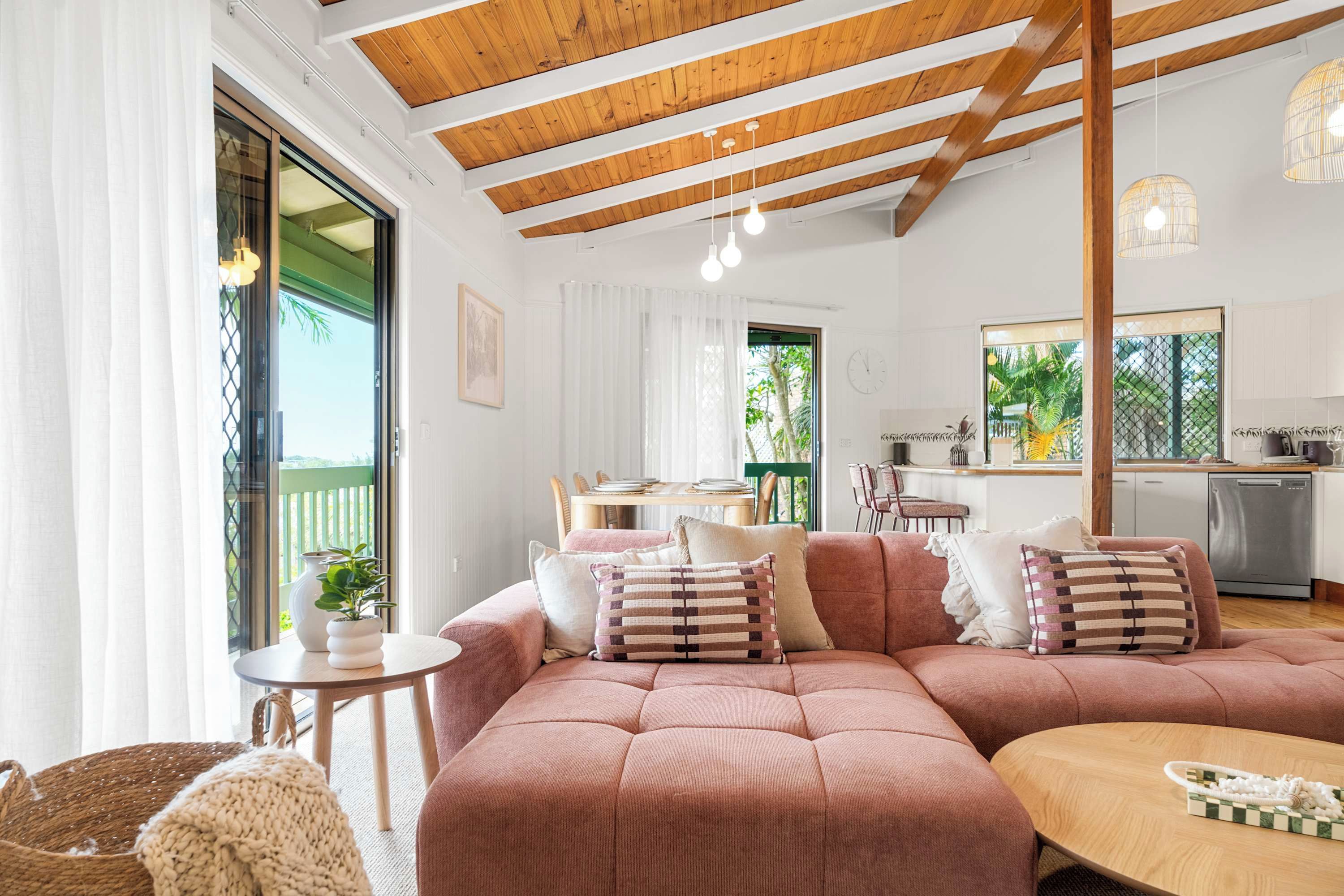 A spacious living area with a large pink sofa, a wooden ceiling, and a view of the kitchen and outdoor area.