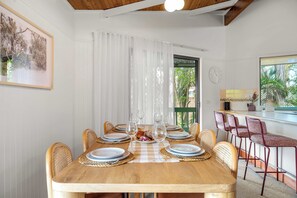 A dining area with a wooden table set for six, surrounded by wicker chairs, a view of greenery outside, and a clock on the wall. - The Breezy Abode - Dog-Friendly, Views & Deck (Caloundra Region)