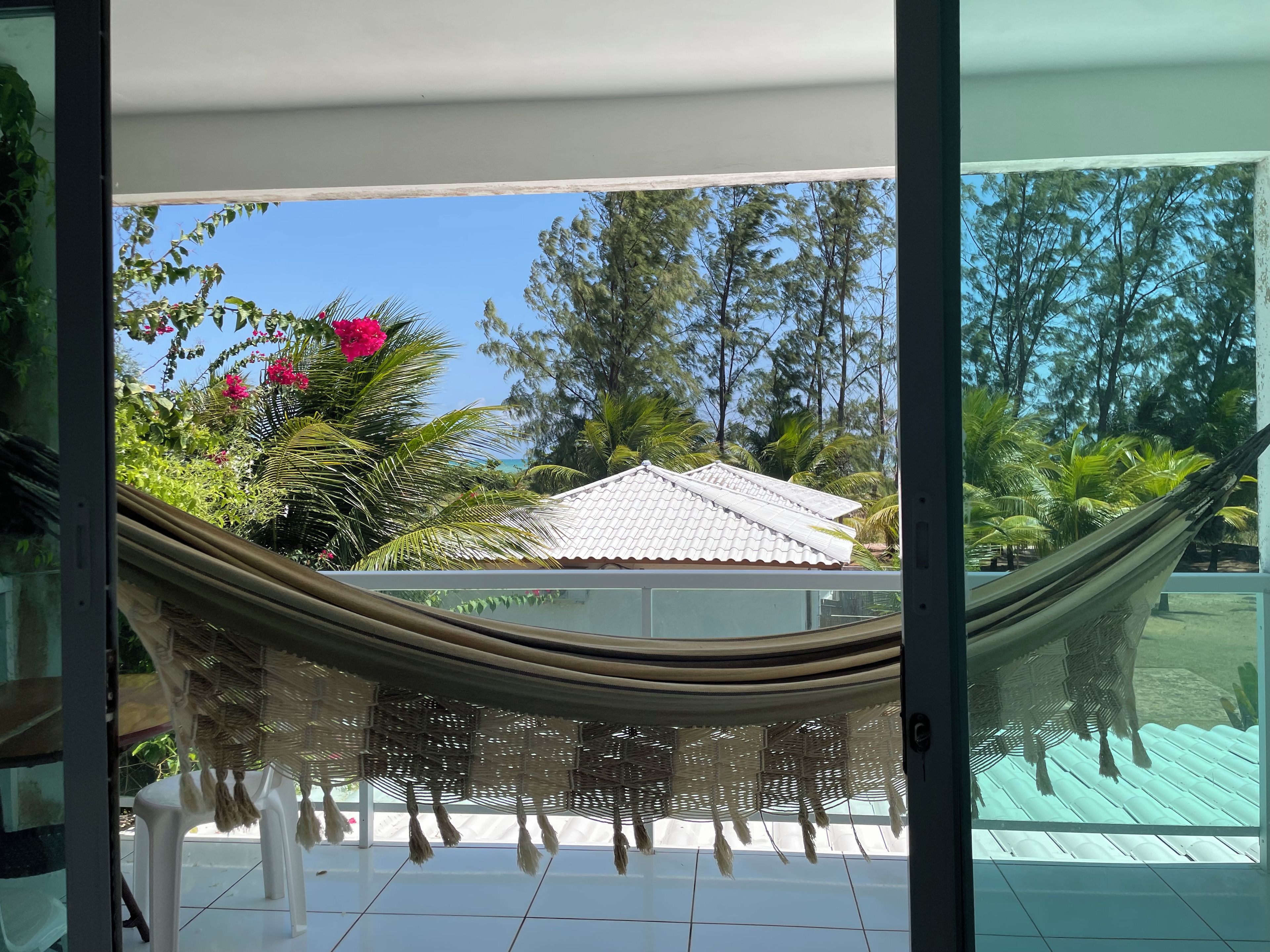 A hammock tied between two windows, a white chair, and a view of a house with a tiled roof and surrounding greenery.