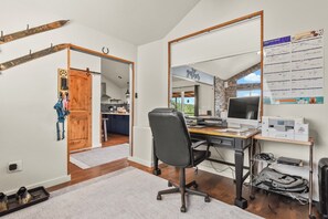 Una oficina en casa con un escritorio grande, computadora e impresora. Hay un espejo en la pared y una puerta que da a una cocina.