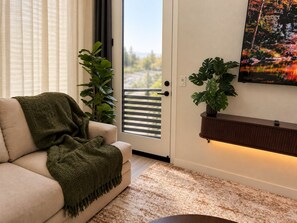 A living room with a sofa, a green blanket, a potted plant, a wall-mounted TV, and a patterned rug. - The Napa Luxe Brand-New 2BR/2BA Sleeps 6. (Napa)