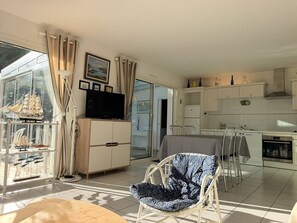 Ein Wohnzimmer mit Fernseher, ein Essbereich mit Stühlen, eine Küche mit Schränken und ein großes Fenster mit Vorhängen.