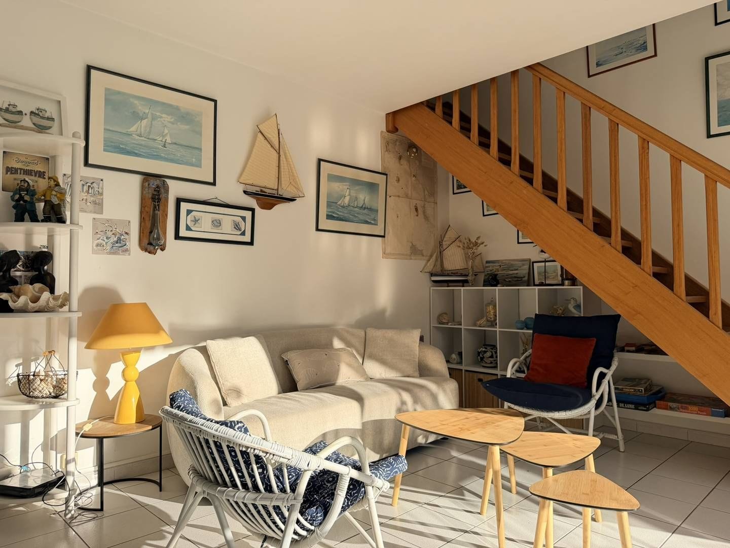 Ein Wohnzimmer mit einer Couch, zwei Sesseln, einem kleinen Tisch, einer Lampe und einem Bücherregal. Im Hintergrund ist eine Holztreppe zu sehen.