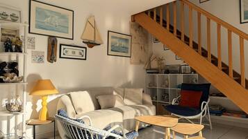 Ein Wohnzimmer mit einer Couch, zwei Sesseln, einem kleinen Tisch, einer Lampe und einem Bücherregal. Im Hintergrund ist eine Holztreppe zu sehen.