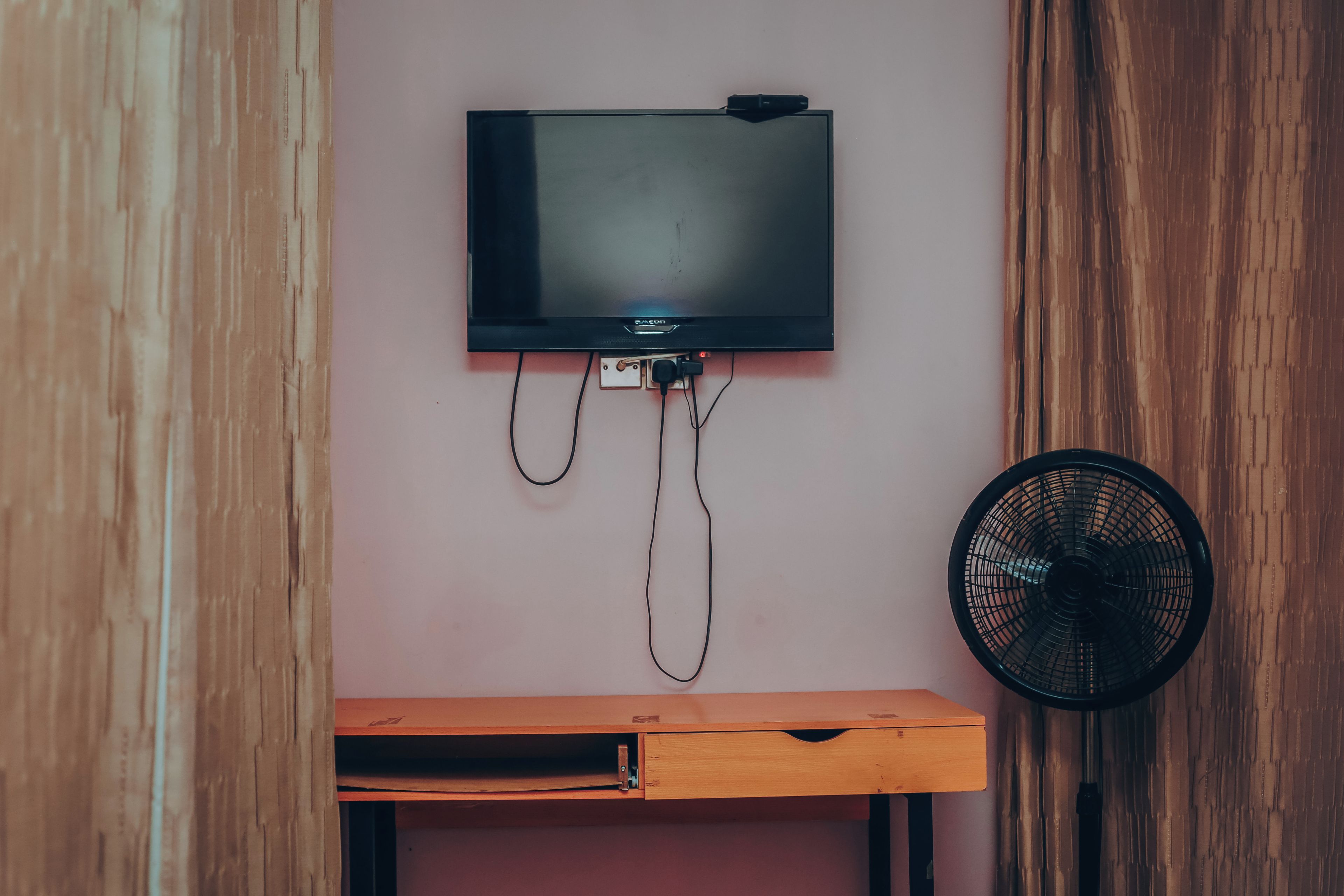 A television mounted on a wall with a wooden desk below and a fan to the right.