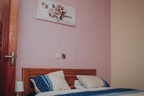 Quarto com cabeceira de madeira, cama com colcha estampada em azul e branco, duas almofadas, abajur e uma obra de arte floral na parede.
