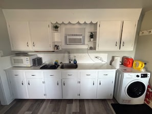 Una cocina compacta con armarios blancos, un microondas, un tostador, un lavamanos y una lavadora.