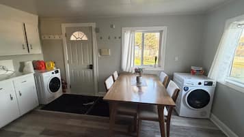 Una cocina compacta con una mesa de madera, dos lavadoras, una puerta con ventana, y un ventanal con vistas al exterior.