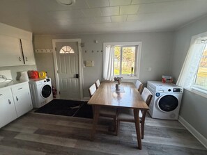 A compact kitchen with a wooden table, two washing machines, a door with a window, and a small window with a view of the outdoors. - Peaceful 2-bedroom bungalow with AC in charming The Pas (The Pas)