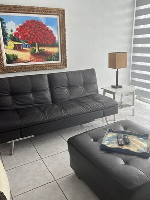 Una sala de estar moderna con un sofá de cuero negro, una mesita auxiliar blanca, una lámpara y un cuadro en la pared.