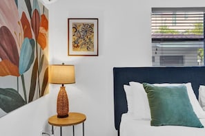 A bedroom with a bed, a bedside table with a lamp, a wall painting, and a window with blinds.