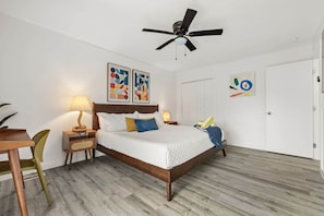 Ein Schlafzimmer mit einem Bett, einem Schreibtisch, einem Stuhl, einem Deckenventilator und Kunstwerken an den Wänden.