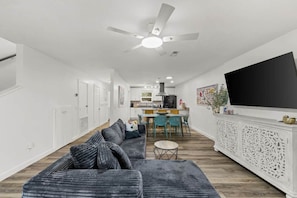 A modern living room with a sofa, dining table, chairs, a TV, and a kitchen in the background. - The Yellow Retreat B | Beach Walk + Private Yard (Panama City Beach)
