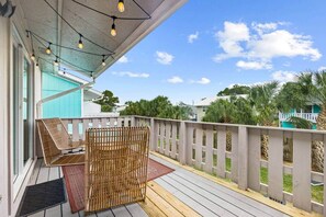 Une terrasse en bois, agrémentée d’une chaise en osier et d’un paravent en bambou, donnant sur un quartier résidentiel avec des palmiers et des maisons.