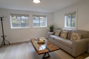 Een woonkamer met een beige bank, een houten salontafel en twee ramen met uitzicht op een tuin.