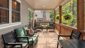 A covered porch with a wooden deck, green cushioned seating, and a stone fireplace. - Covewood by Avantstay Lakefront w/ Private Shoreline Kayaks + Paddle Boards + Firepit (Lakeville)