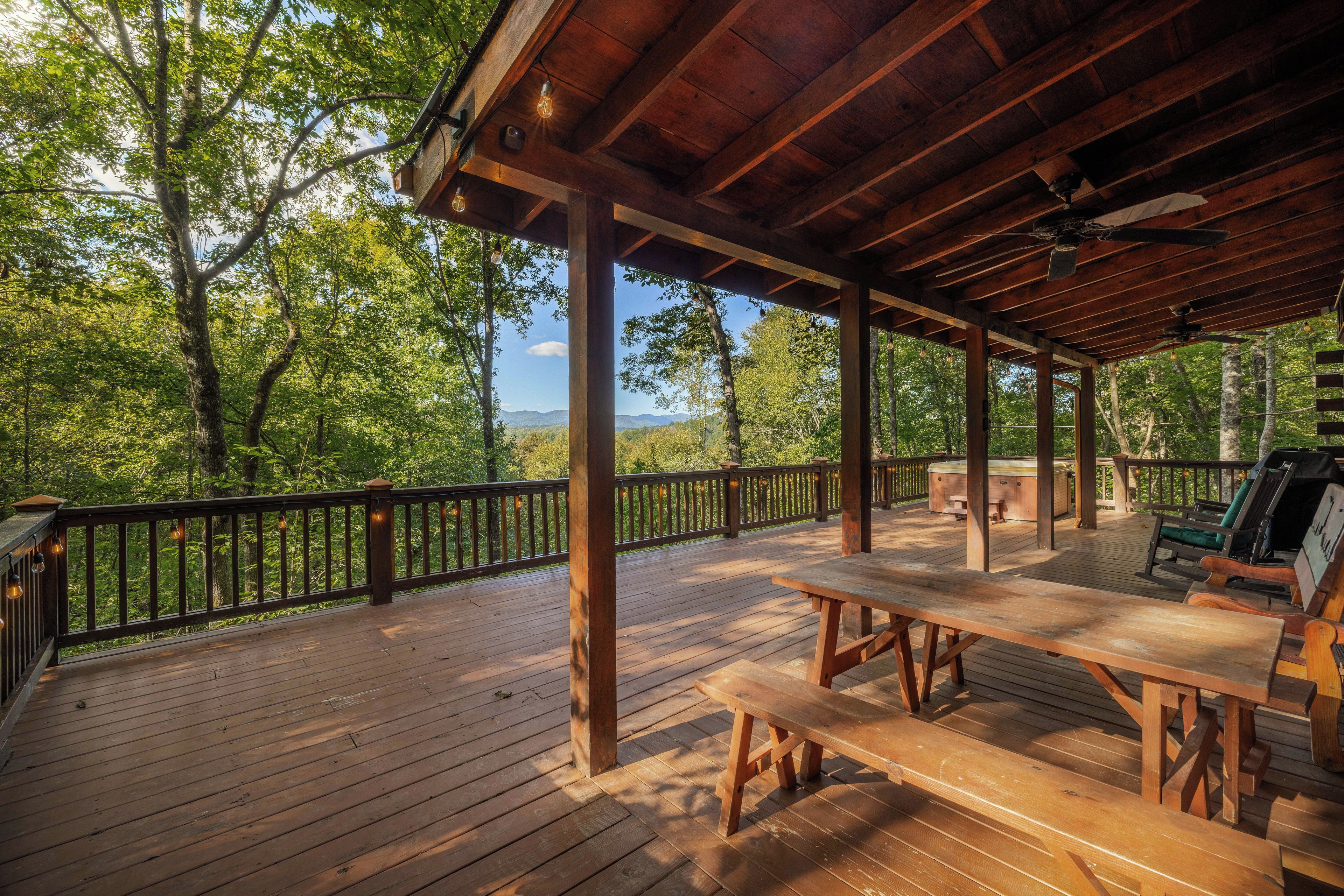 A wooden deck with picnic tables and a hot tub, surrounded by trees and a view of the mountains.