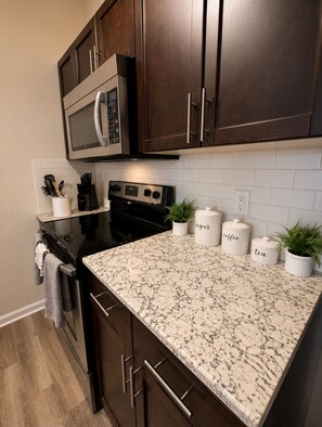A kitchen with dark wood cabinets, a stainless steel microwave, a black stove, and a marble countertop. There are three white containers labeled 'sugar,' 'coffee,' and 'tea.' - Stylish 2BR King Suite Near Uptown (Charlotte)