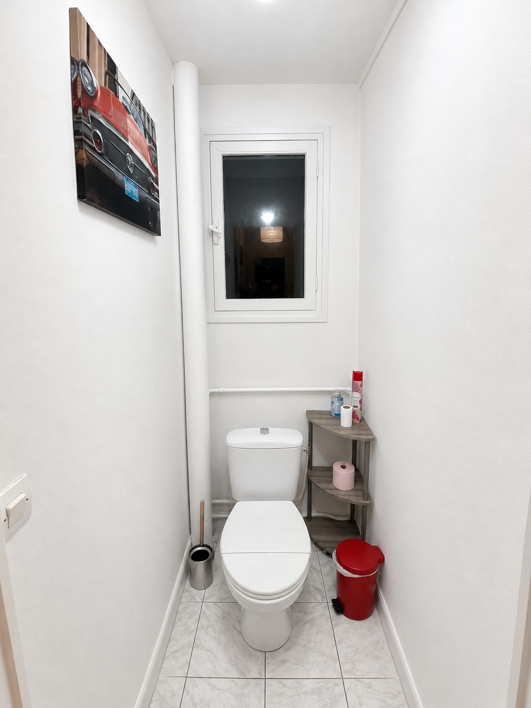 Et lille badeværelse med et hvidt toilet, et lille vindue, en hylde med toiletartikler og en rød spand.