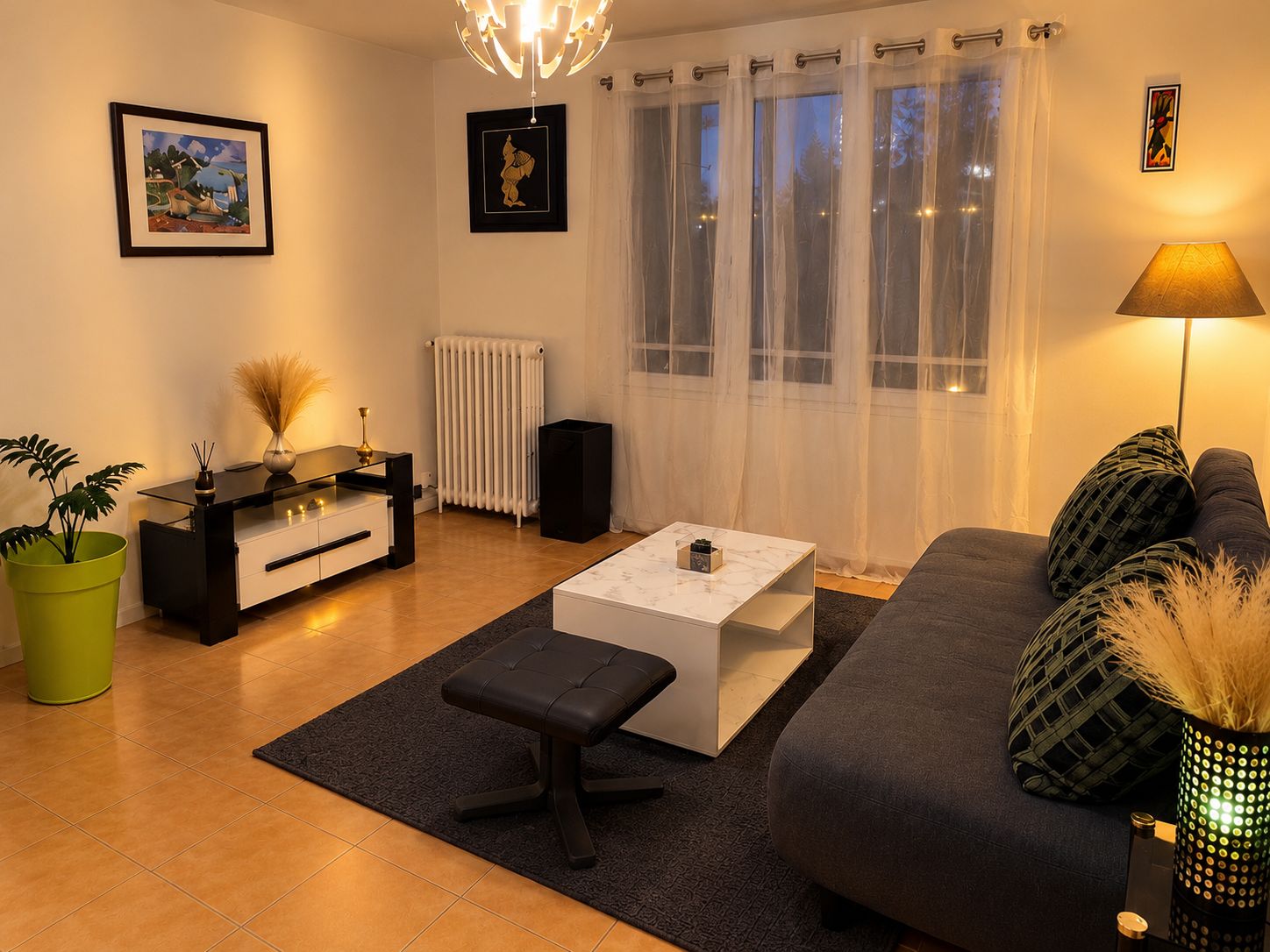 Ett modernt vardagsrum med en soffa, ett soffbord och ett sidobord med en lampa. Det finns ett element, en krukväxt och konstverk på väggarna.