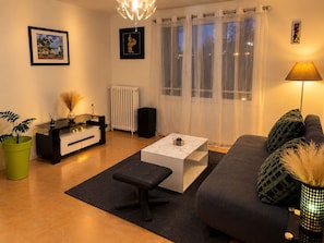 Ein modernes Wohnzimmer mit einer Couch, einem Couchtisch und einem Beistelltisch mit Lampe. Es gibt eine Heizung, eine Zimmerpflanze und Kunstwerke an den Wänden.
