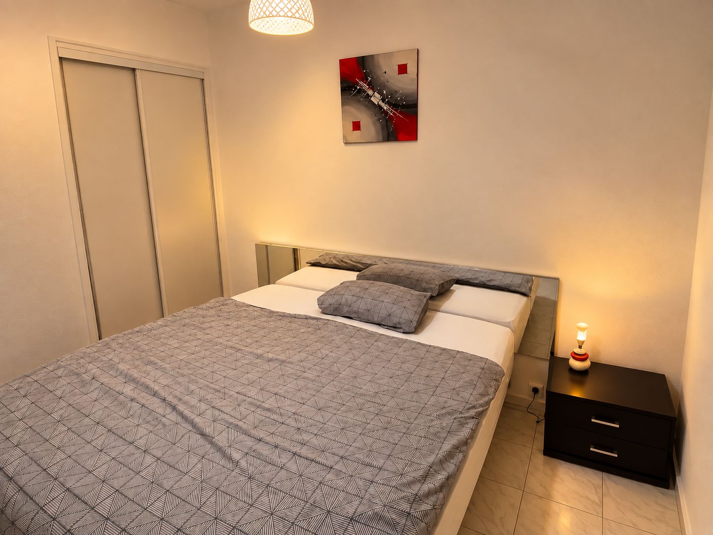 Ein Schlafzimmer mit Bett, Kissen, einem Nachttisch, einer Lampe und einem Bild an der Wand.