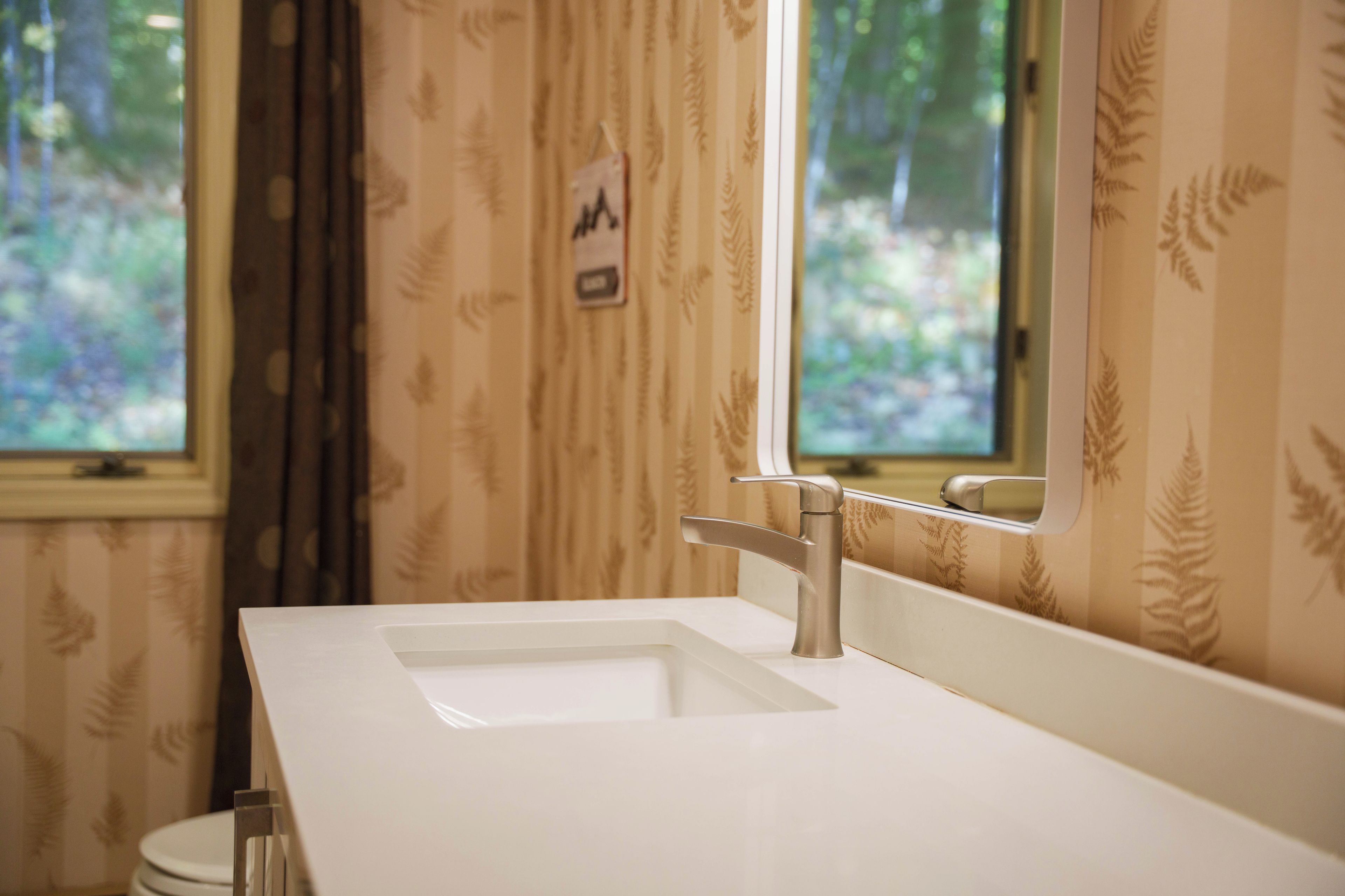 A bathroom with a white sink, a mirror, and a window with a view of greenery.