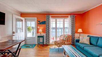 Ein Zimmer mit einem Holztisch, einem blauen Sofa, einem Korbstuhl und Blick durch ein großes Fenster.