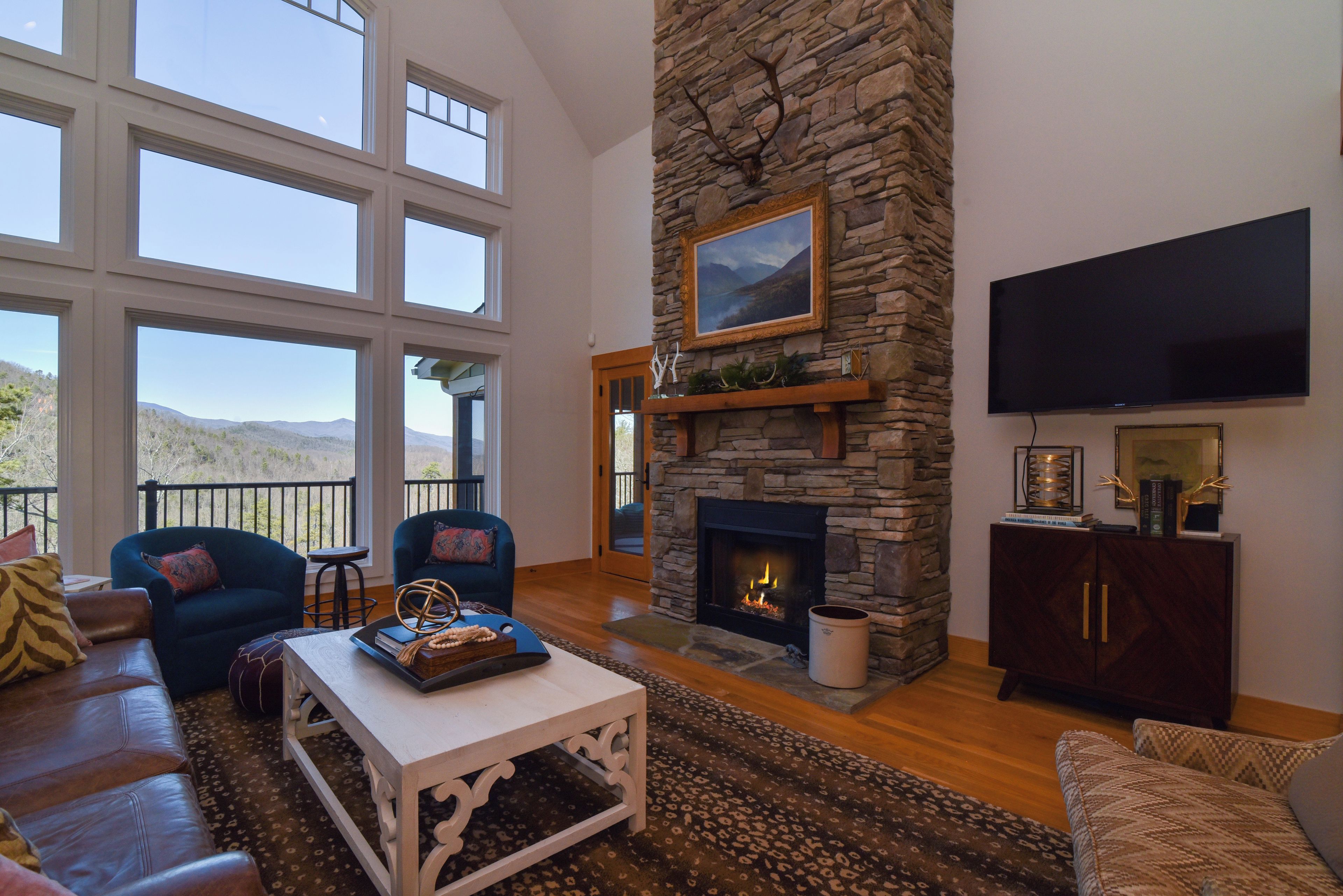 A spacious living room with a stone fireplace, large windows, and a flat-screen TV.