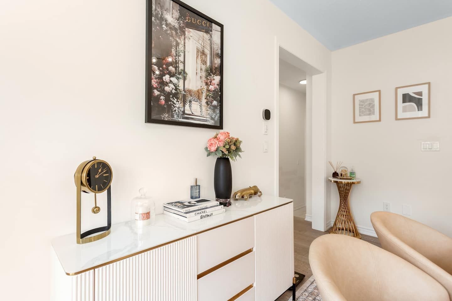 A modern interior with a white console table, a clock, books, a vase with flowers, and a framed Gucci artwork on the wall.