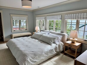 A bedroom with a large bed, bedside tables, a bench, and a view of greenery through the windows. - The Choo Choo House- Luxe renovated home w/ ADU, Sleeps 10, AC, Wrap-around Deck (Livingston)