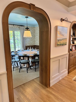 An interior view of a dining area with a wooden archway leading to a table set for a meal, a hanging light fixture, and a framed landscape painting on the wall. - The Choo Choo House- Luxe renovated home w/ ADU, Sleeps 10, AC, Wrap-around Deck (Livingston)