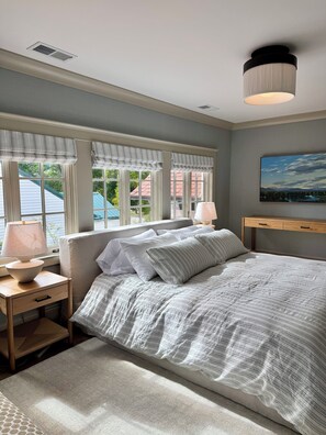 Un dormitorio con cama, mesitas de noche, lámpara, un cuadro en la pared y ventana con cortinas.
