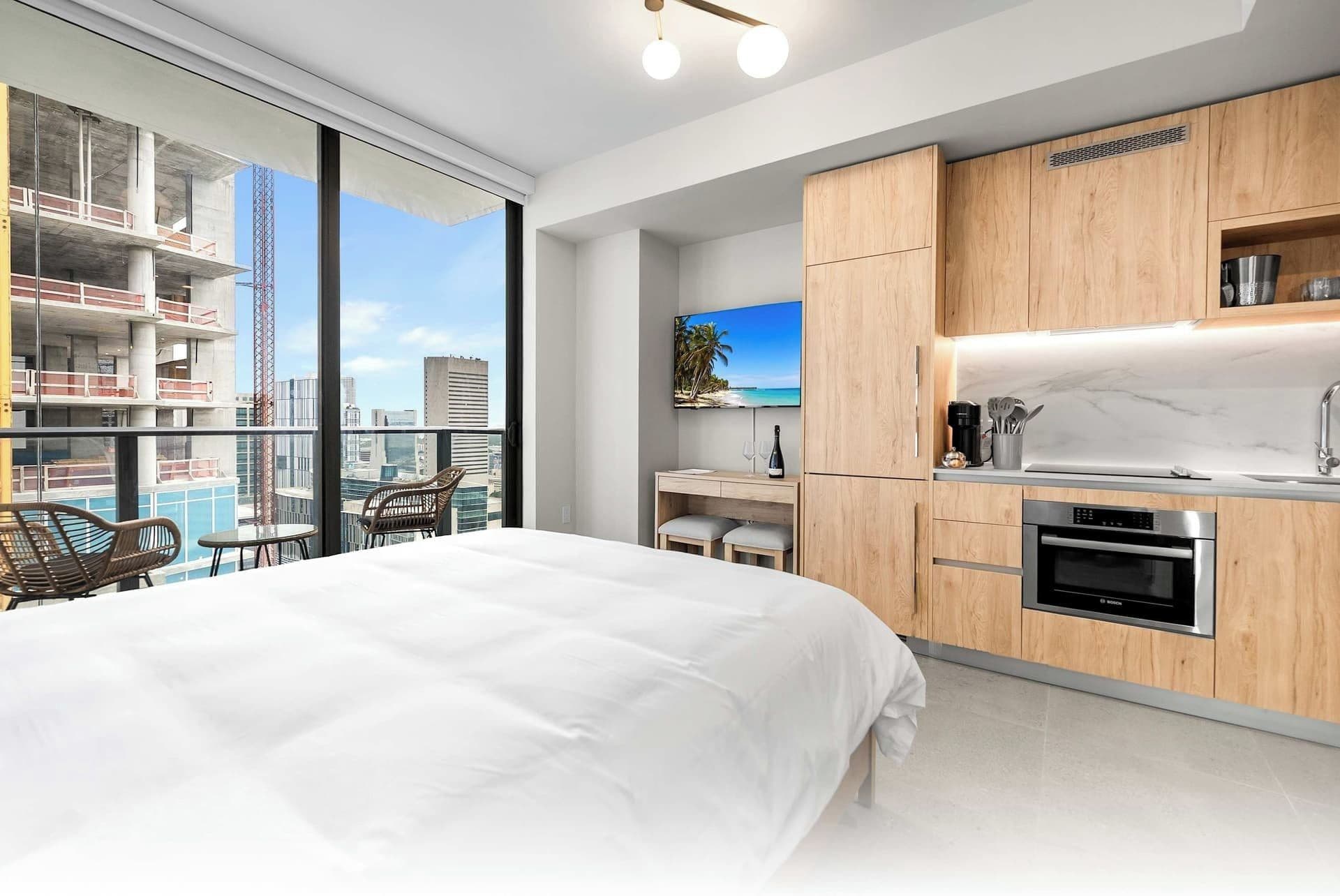 A modern bedroom with a large bed, a desk, and a view of the city through floor-to-ceiling windows.
