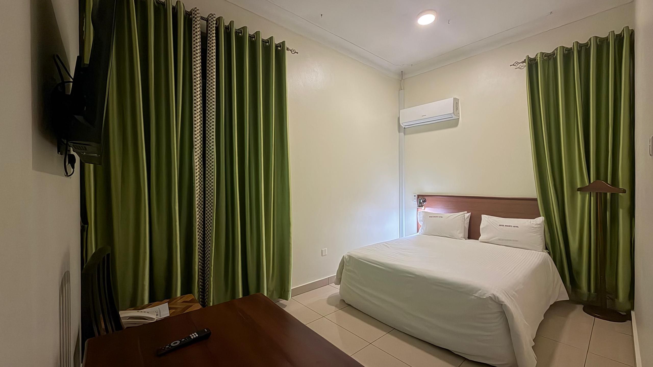 A hotel room with a bed, a TV mounted on the wall, a table with a remote control, and green curtains.
