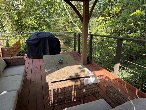 A wooden deck with outdoor furniture, including a sofa and a table, surrounded by lush greenery.
