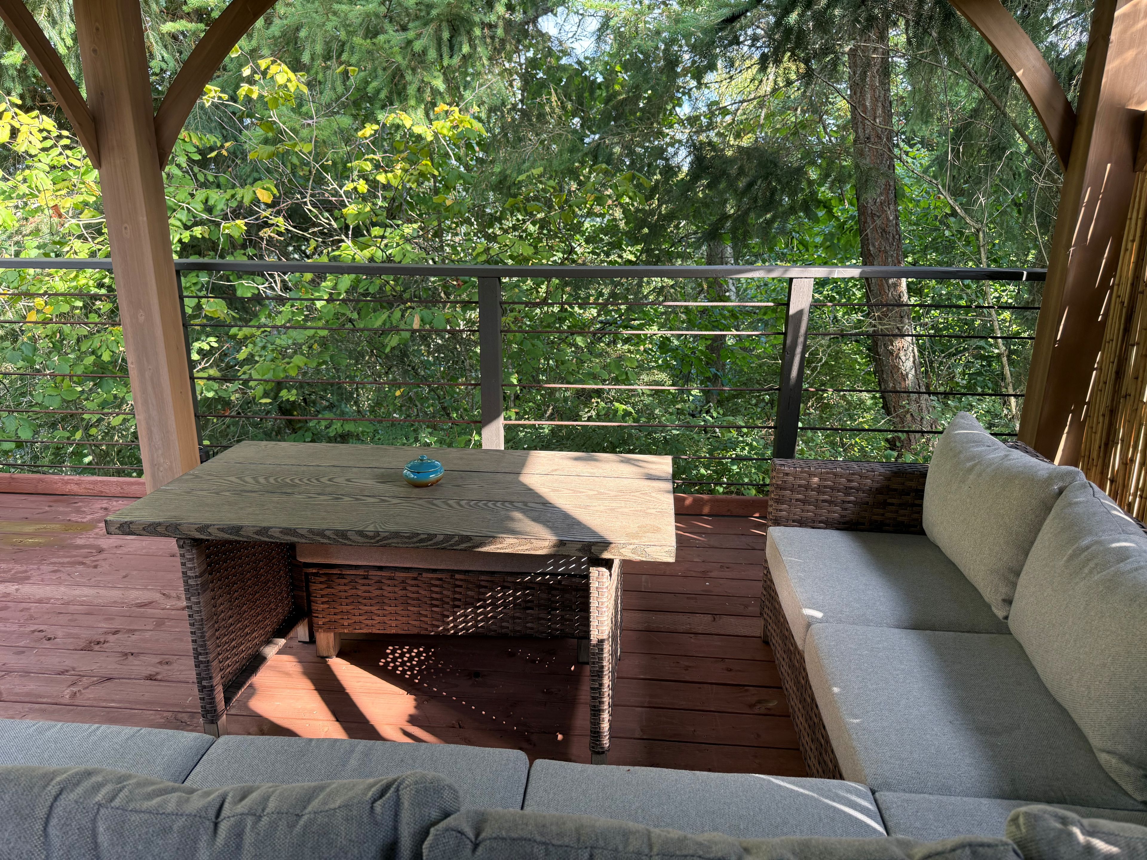 Un patio al aire libre con mesa de madera, mobiliario de mimbre y vistas a zona boscosa.
