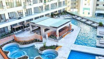 A modern hotel with multiple swimming pools, a covered seating area, and a view of adjacent high-rise buildings.