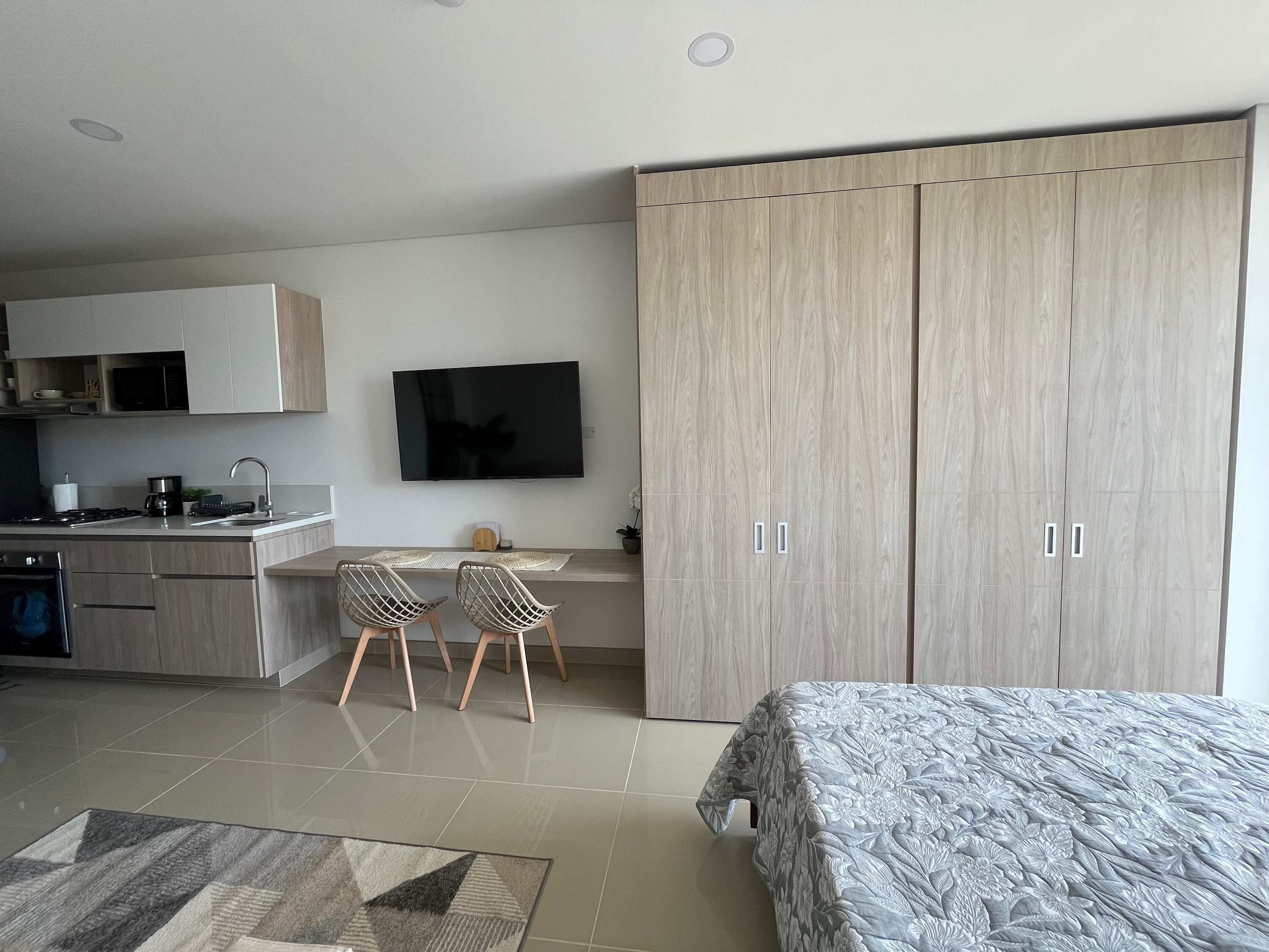 A modern bedroom with a bed, a TV mounted on the wall, a kitchenette with cabinets, and a large wardrobe.