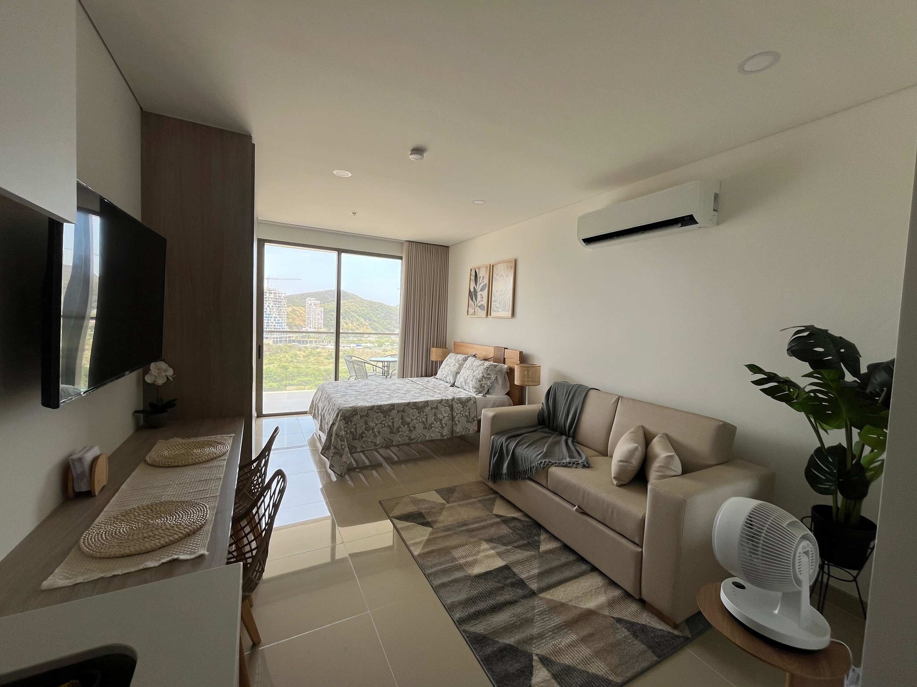 A modern hotel room with a bed, a sofa, a TV, and a balcony with a view of a cityscape.