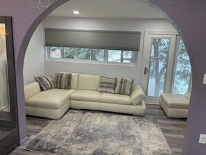 A living room with a beige sectional sofa, a patterned rug, and a window with blinds. - Soothing 1-bedroom studio with AC in charming Guttenberg (Guttenberg)