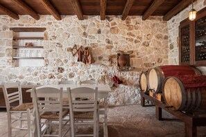 A stone-walled room with wooden beams, a table with chairs, wine barrels, and shelves with bottles. - VILLA LITHOS, Your Villa (Chania)
